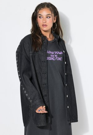 Jeune femme avec des boucles d'oreilles créoles porte une veste en jean noire oversize par-dessus un t-shirt avec l'inscription « Slow down you're doing fine » en texte violet.