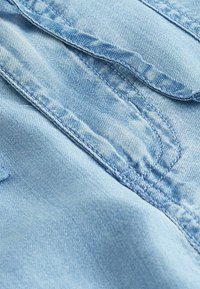 Tissu en denim bleu clair avec une texture lisse, des coutures visibles et un bord replié, mettant en valeur les détails de construction du vêtement.