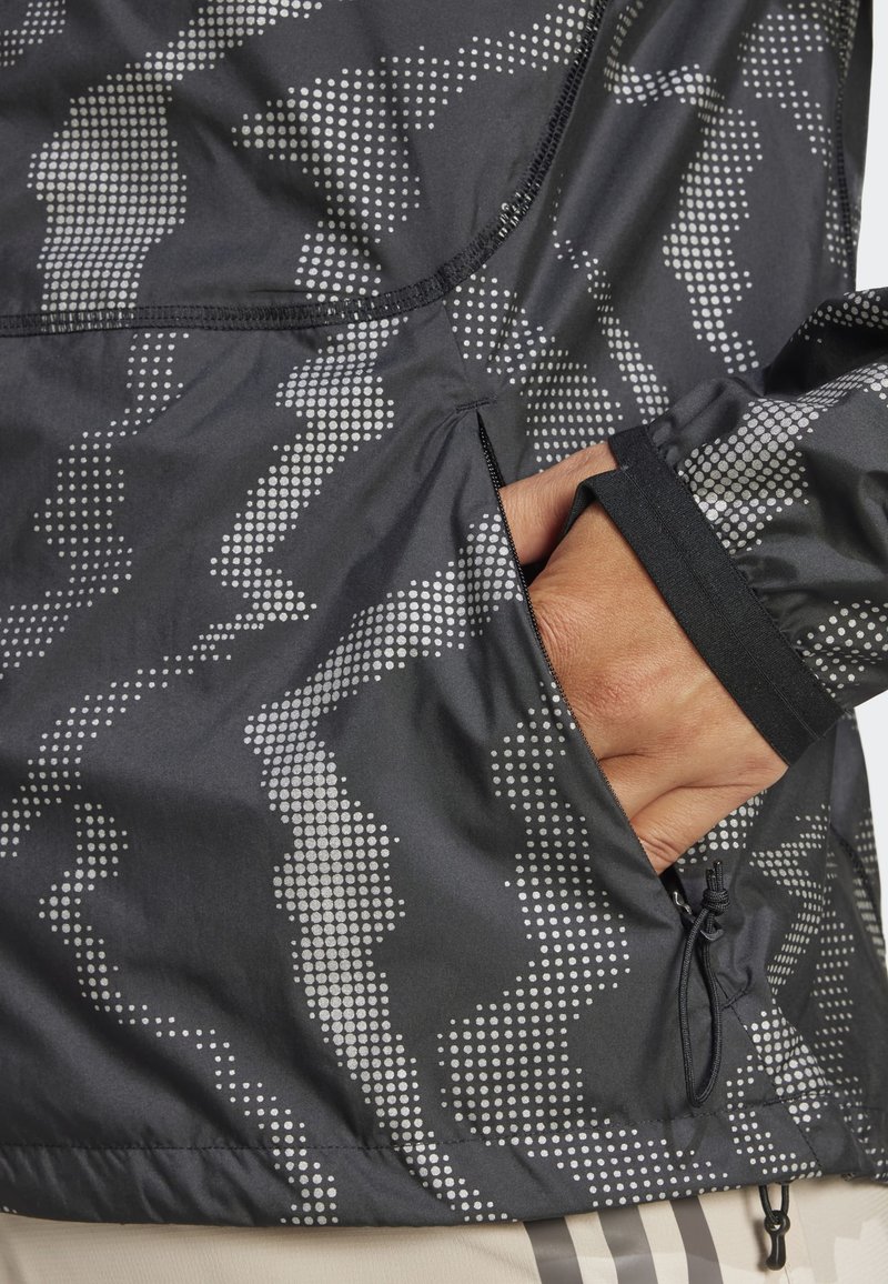 Schwarze Jacke mit Mustern, die ein silbernes Punktdesign aufweist, mit Reißverschlusstasche und verstellbarem Kordelzug am Saum. Weicher, leichter Stoff.