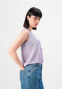 Ung jente med mørk bob-frisyre som har på seg en ermeløs lavendelstrikket topp og blå jeans, står sidelengs med hendene i lommene.