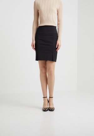 Femme portant un haut beige côtelé à manches longues, une jupe crayon noire taille haute et des talons noirs à bride à la cheville avec des clous dorés, debout sur un sol blanc.