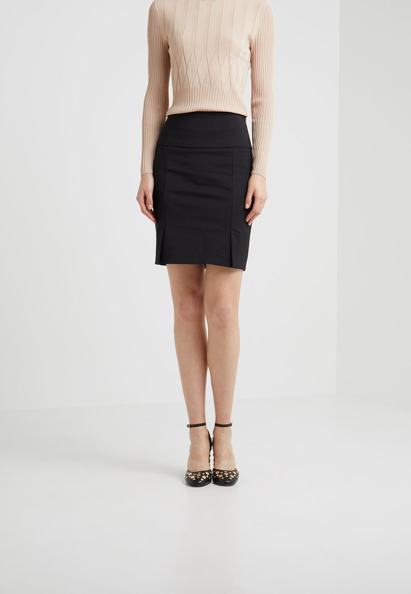 Femme portant un haut beige côtelé à manches longues, une jupe crayon noire taille haute et des talons noirs à bride à la cheville avec des clous dorés, debout sur un sol blanc.