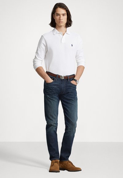 Witte langarm polo, donkerblauwe jeans, leren bruine riem en tan kleurige suède schoenen. Model staat met handen in de zakken tegen een lichte achtergrond.