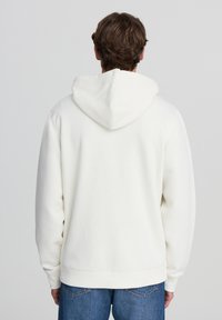 Felpa bianca con cappuccio e trama morbida, dotata di tasca a canguro e polsini a costine, abbinata a jeans blu. Vista posteriore mostrata.