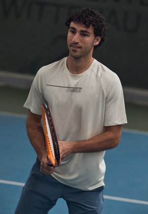 Hellgraues Sport-T-Shirt mit kurzen Ärmeln und einem Logo, kombiniert mit dunklen Shorts. Die Person hält einen Tennisschläger mit einem orangefarbenen Rand.
