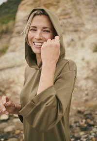 Sweat à capuche vert olive à coupe décontractée, manches trois-quarts, et texture lisse, porté par une femme en extérieur.