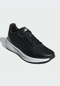Zapatilla deportiva negra con parte superior de tela texturizada, cordones, diseño de tres rayas y una suela blanca contrastante con un patrón de olas.