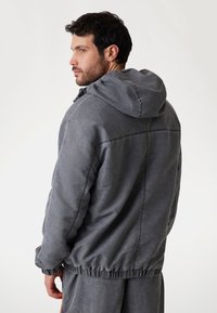Chaqueta gris con capucha, con dobladillo elástico y un corte casual, que presenta costuras visibles y una textura suave. Ideal para atuendos en capas.