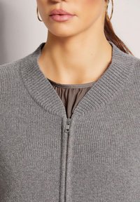 Grau gerippte Reißverschlussjacke mit einem bündchenbesetzten Kragen und glattem Reißverschluss, über einer braunen Bluse mit gerafft geschnittenem Ausschnitt getragen.