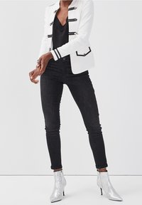 Vit jacka med svarta detaljer och knappdetaljer; svarta tights jeans; blanka silverfärgade ankelboots med spetsiga tår; släta texturer.