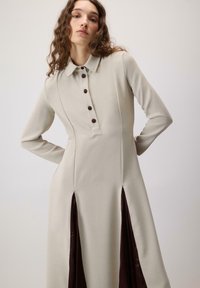 Langærmet kjole i lys beige stof med krave, prydet med brune knapper og slidser i siderne, der afslører mørkebrune satin detaljer.