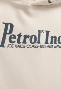 Tissu beige avec texte brodé en gris foncé : « Petrol Ind - Ice Race Class - Big Lake » avec un motif de drapeau à damier partiel.