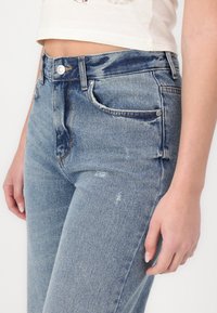 Světle modré džínové kalhoty s vysokým pasem, distressed detaily a klasickým pětikapsovým designem. Obsahují kovový knoflík a zipové zapínání.