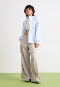 Persona che indossa una giacca leggera blu chiaro con zip, pantaloni beige a gamba larga, top corto, scarpe argento e occhiali trasparenti, in piedi su un pavimento verde.
