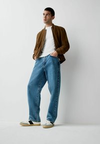 Brūna zamšādas bumbvedēja jaka virs baltas T-krekla, kopā ar gaiši zilām brīvām džinsa biksēm un daudzkrāsainām sporta kurpēm.