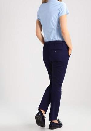 Personne portant une chemise à manches courtes bleu clair et un pantalon bleu marine, les mains dans les poches, debout devant un fond blanc uni.