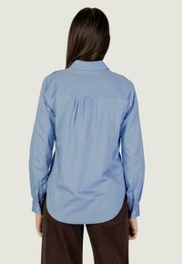 Persona che indossa una camicia a maniche lunghe di colore azzurro chiaro con dettaglio arricciato sulla schiena e pantaloni scuri, in piedi e di spalle su uno sfondo neutro.