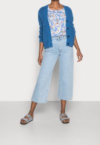 Blauer gestrickter Cardigan mit Knöpfen über einem blumigen Oberteil, kombiniert mit lässiger heller blauer Jeans und grauen Sandalen. Sauberes, modernes Erscheinungsbild.