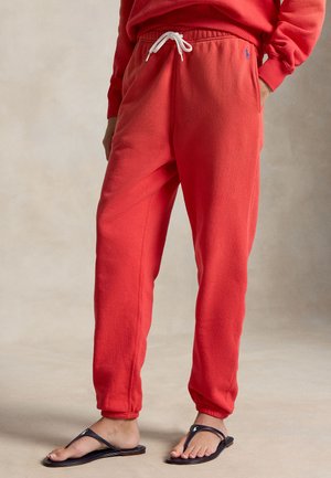 Osoba ubrana w jasnoczerwone spodnie dresowe ze sznurkiem w kolorze białym, w czarnych klapkach, stojąca na neutralnym tle.