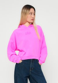 Rosa cropped hettegenser med hette og frontlomme, som har en liten "GAP" logo i midten. Kombinert med mørkeblå denimjeans.