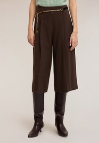 Culottes plissées marron avec ceinture chaîne dorée, portées avec de grandes bottes en cuir marron brillant. Texture douce du tissu visible.