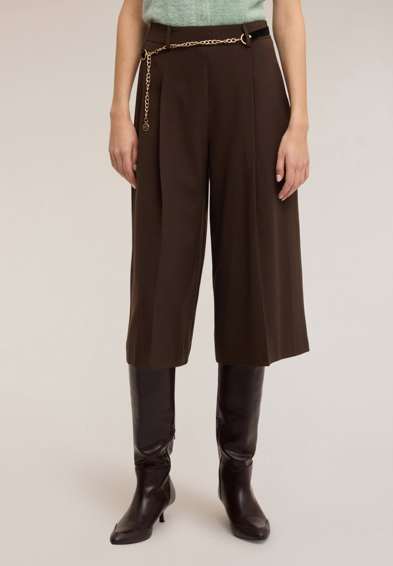 Culottes plissées marron avec ceinture chaîne dorée, portées avec de grandes bottes en cuir marron brillant. Texture douce du tissu visible.