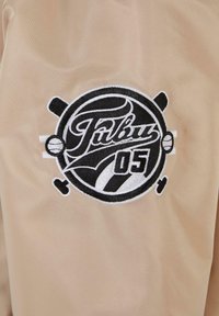 Beige kangas, millel on musta ja valgega tikitud logo "Päulus 05" ja dekoratiivsed elemendid, mis meenutavad spordivahendeid.