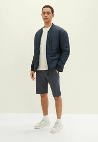 Marineblau gesteppte Bomberjacke mit Reißverschlusstasche, über einem hellen T-Shirt und gemusterten Shorts getragen. Das Model trägt weiße Sneaker und Socken.