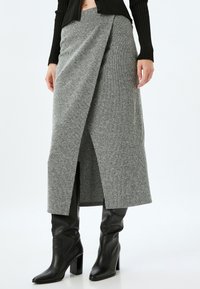 Jupe asymétrique grise en maille côtelée, fabriquée en tissu tricoté texturé, avec une fente à l'avant et associée à des bottes noires montantes.