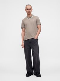 Homme debout, une main dans la poche, portant un polo beige, un jean noir ample et des chaussures noires sur un fond clair uni.