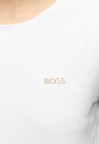 Camiseta blanca con texto bordado en beige "BOSS" en el pecho, llevada por una persona con tono de piel claro.