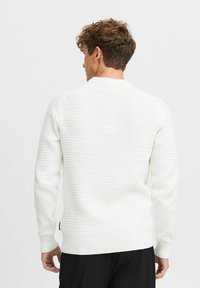 Pull en tricot blanc avec un motif texturé, des poignets et un ourlet côtelés, un col rond et une petite étiquette logo en bas à gauche.