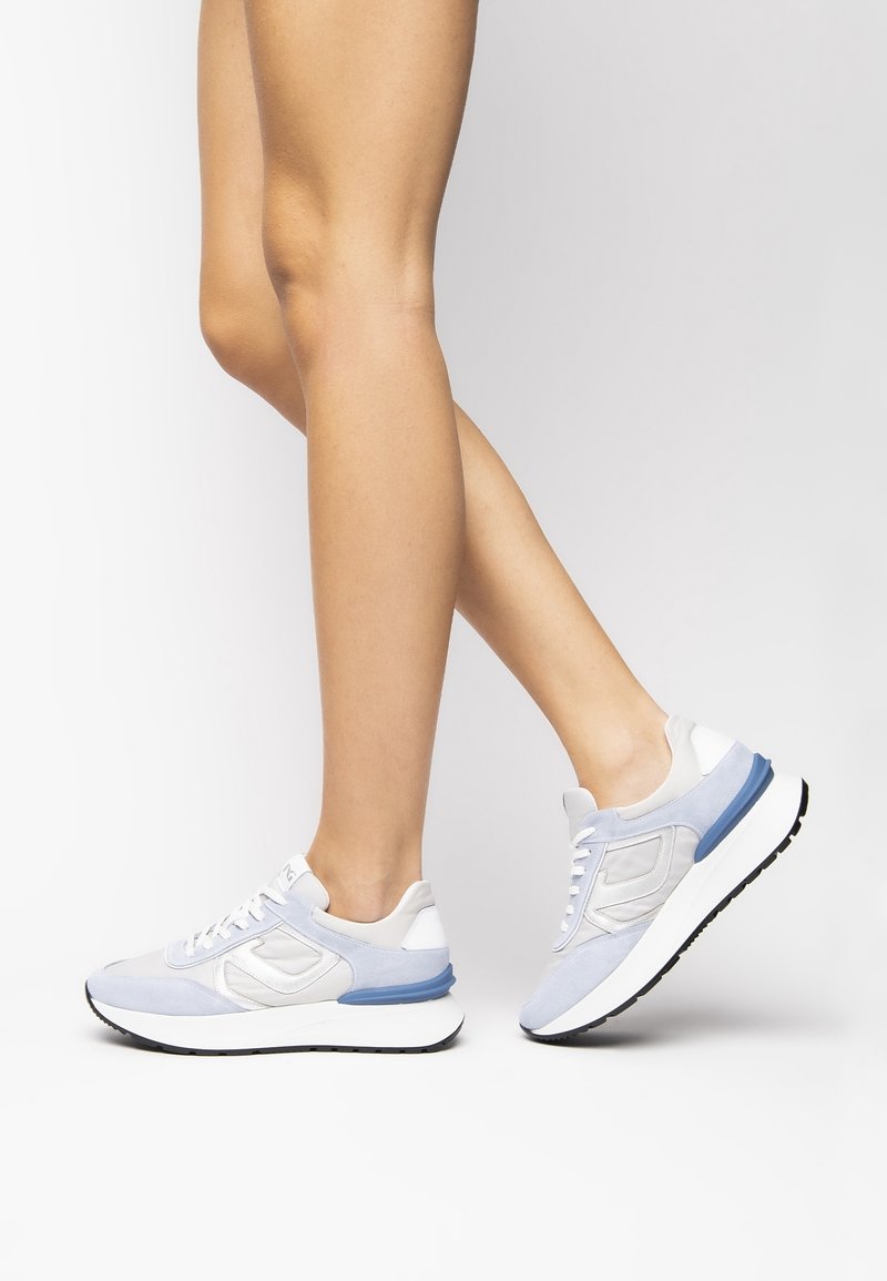 Jambes portant des baskets blanches et bleu clair avec des lacets blancs, un pied posé à plat sur le sol, l'autre levé sur la pointe des pieds sur fond gris.
