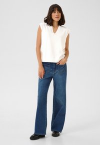 Haut tricot sans manches blanc avec un col en V, associé à un jean en denim large bleu clair. Des chaussures noires à semelles épaisses complètent le look.