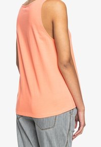 Top sin mangas en color coral, hecho de algodón suave, con un corte relajado, escote redondo y una textura suave, combinado con jeans de color gris claro.