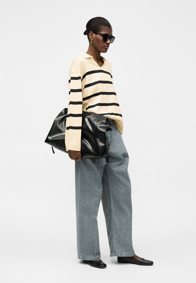Γυναίκα που φοράει πουλόβερ με ρίγες, τζιν με φαρδιά πόδια, μαύρα μπαλαρίνια, γυαλιά ηλίου και κρατά μια μεγάλη μαύρη δερμάτινη τσάντα σε απλό φόντο.
