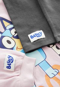 Zwei Baumwoll-T-Shirts: eines grau, eines rosa, mit "Bluey"-Labels; das rosa Shirt zeigt ein buntes Cartoon-Hundedesign auf einem hellen Hintergrund.