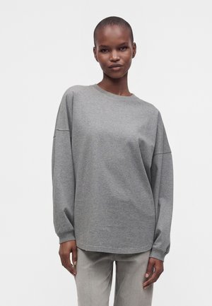 Femme aux cheveux courts portant une chemise grise ample à manches longues et un pantalon gris clair sur un fond blanc uni.