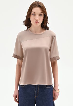 Jeune femme aux cheveux bruns ondulés portant un haut en satin beige à manches courtes et un jean bleu foncé, debout devant un fond clair uni.