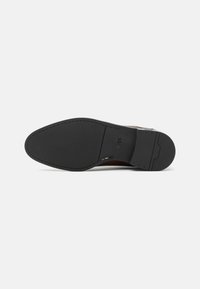 Tălpi din piele maro cu o bază din cauciuc negru, având un model texturat și un brand gravat pe lateral. Formă rotundă la vârf.