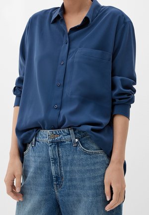 Personne portant une chemise ample bleu marine à boutons avec les manches retroussées, rentrée dans un jean en denim bleu taille haute.