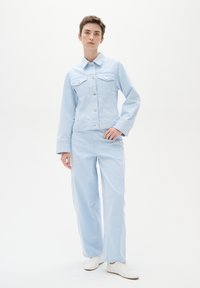 Gai zila denima jaka ar divām krūšu kabatām, piecu pogu aizdare un atbilstošas gaiši zilas bikses. Modelis valkā baltas sporta kurpes.