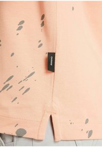 Pfirsichfarbener Baumwoll-T-Shirt mit grauen Spritzmustern, seitlichen Schlitzern am Saum und einem schwarzen Etikett mit dem Text "CHASIN'".