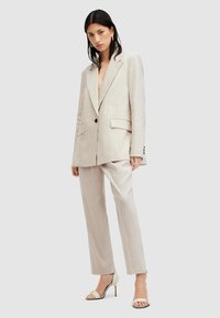Vrouw in een beige oversized blazer en bijpassende broek, staand met een neutrale uitdrukking tegen een effen achtergrond.