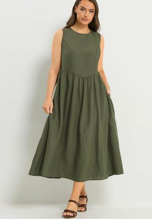 Kobieta stoi w oliwkowej, bezrękawnej sukience do kolan z dopasowanym gorsetem i marszczonym dołem, połączonej z brązowymi sandałami na pasku wokół kostki.
