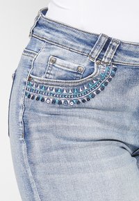 Jean en denim bleu clair avec des embellissements décoratifs bleus et argentés sur la poche avant. Dispose d'un design classique à cinq poches.