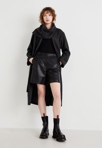 Svart ullkåpe med stor krage, kombinert med svarte skinnshorts og kraftige svarte ankelstøvler med gule detaljer.