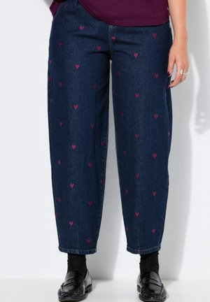 Pantalones vaqueros azul oscuro con pequeños patrones de corazones rosas, usados con calcetines negros y mocasines negros por una persona que está de pie contra un fondo blanco.