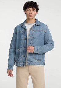 Chaqueta de mezclilla azul claro con cuello, botones frontales, bolsillos en el pecho y un corte relajado, llevada sobre una camiseta gris.