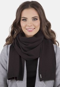 Mørkebrunt strikket tørklæde med blød tekstur, lagt om halsen, med glatte kanter og et subtilt mærke i enden.